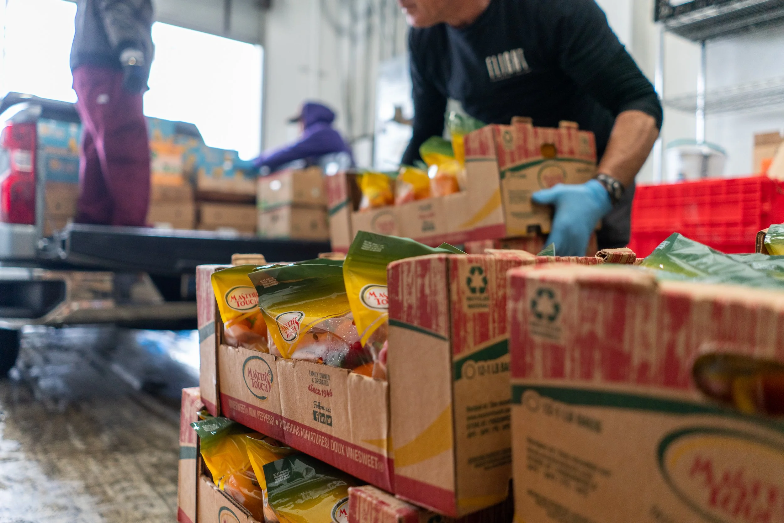 Multiple boxes of mixed fruit packages being packed or sorted by workers in a warehouse or grocery store, with one worker in gloves handling the boxes, and others in the background.