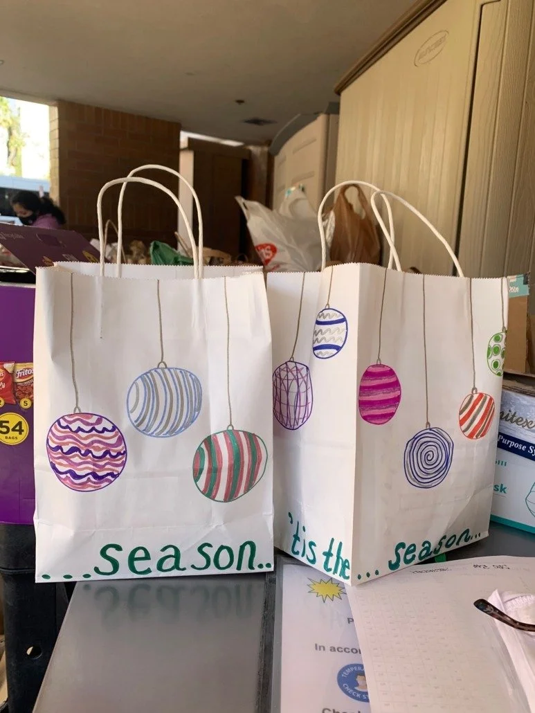 Two white shopping bags decorated with colorful, hand-drawn ornaments hanging from thin lines. The bags have the phrase "...season... it's the..." written in green letters at the bottom.