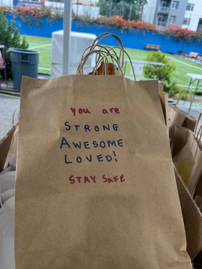 Brown paper bag with a handwritten message: 'You are strong, awesome, loved! Stay safe' in red and blue ink. Background includes greenery, buildings, and outdoor furniture.