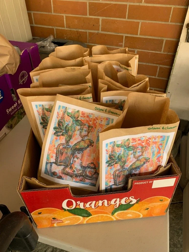 A box of paper lunch bags with colorful artwork and labels, labeled 'salami & cheese'. The box is labeled 'Oranges' and features images of oranges, set on a table against a brick wall.