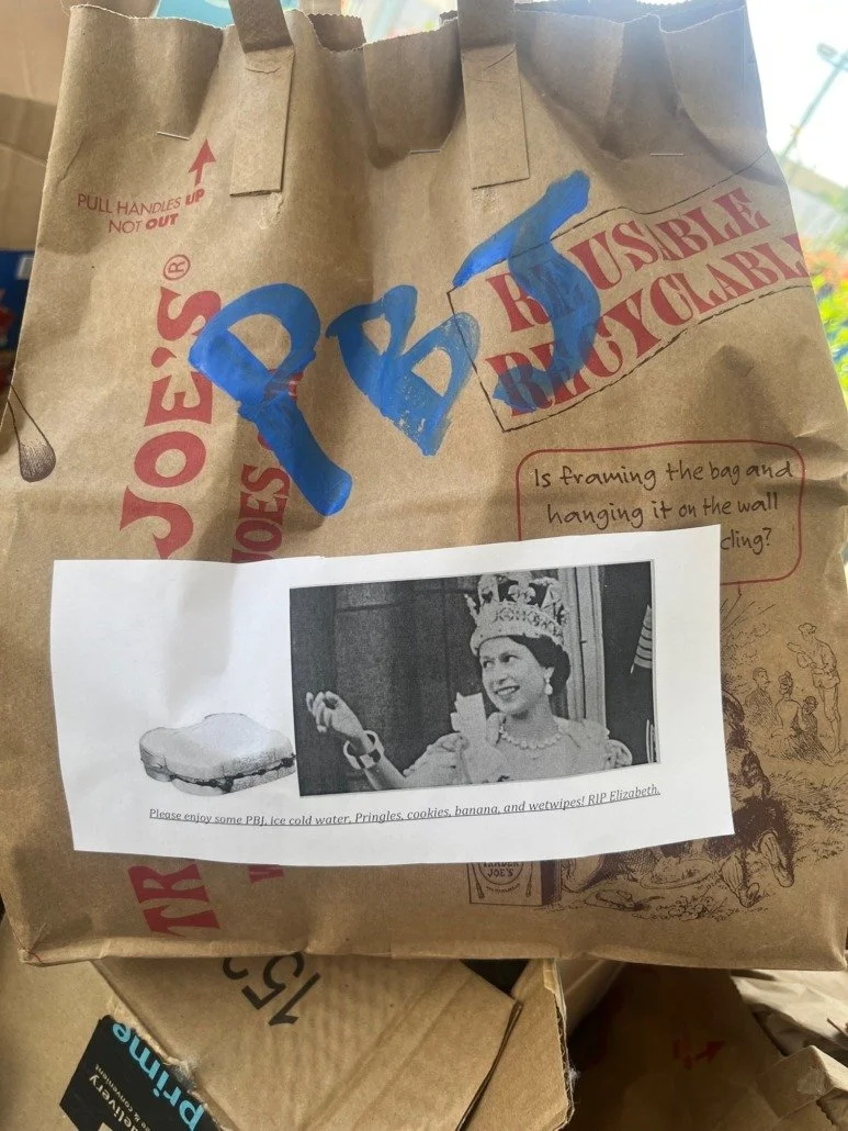 A paper bag from Joe's that has blue graffiti letters 'RBS' painted on it. There is a printed black-and-white photo of Queen Elizabeth II holding a glass and smiling, with a caption beneath that says, 'Please enjoy some PBJ, ice cold water, Pringles,