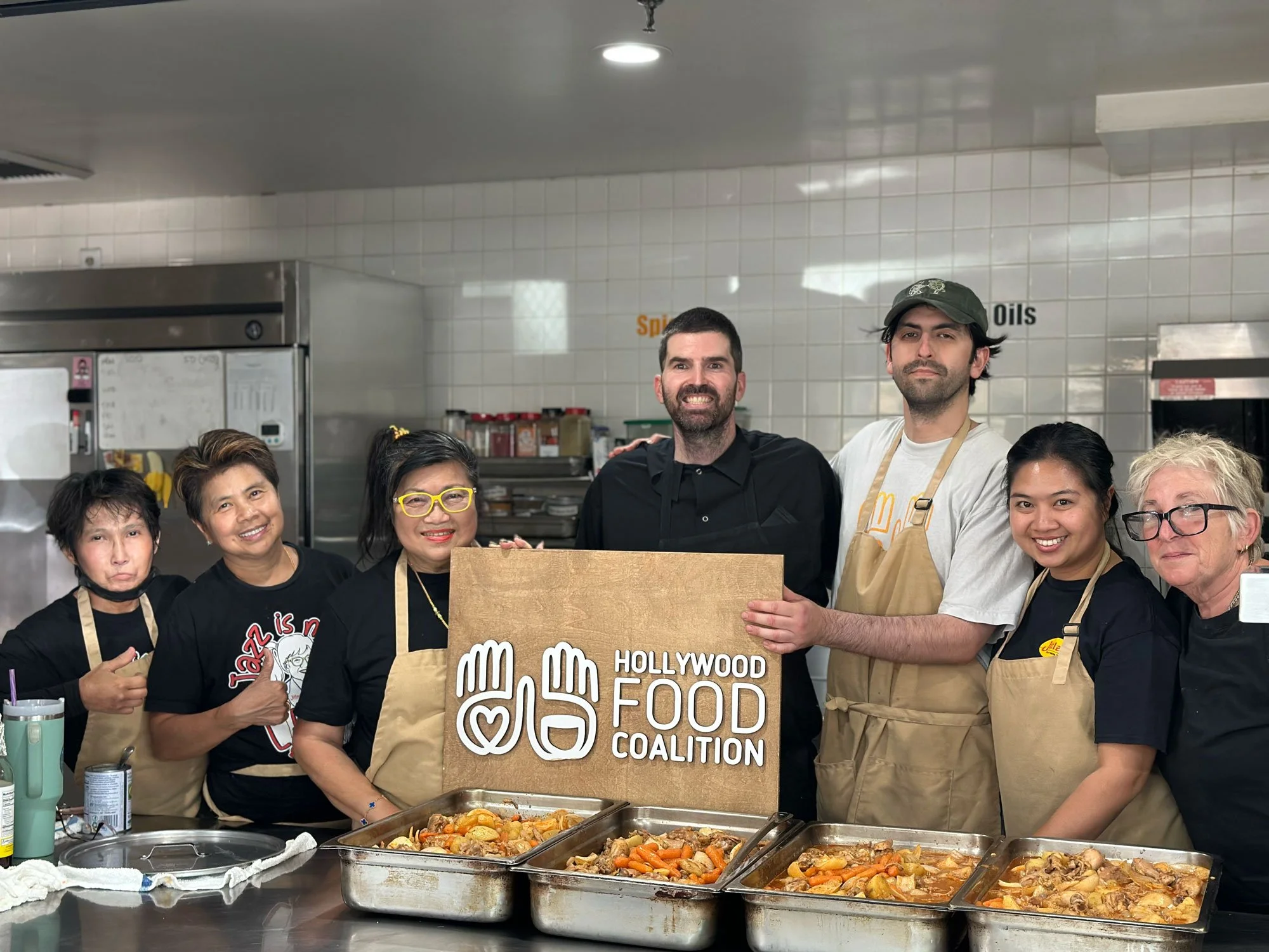 Group of people in a kitchen holding a sign that says Hollywood Food Coalition, with trays of food in front of them.