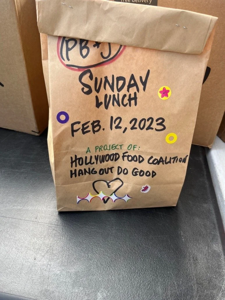Brown paper bag with handwritten text about a Sunday lunch event on February 12, 2023, organized by Hollywood Food Coalition, decorated with colorful stickers and drawings.
