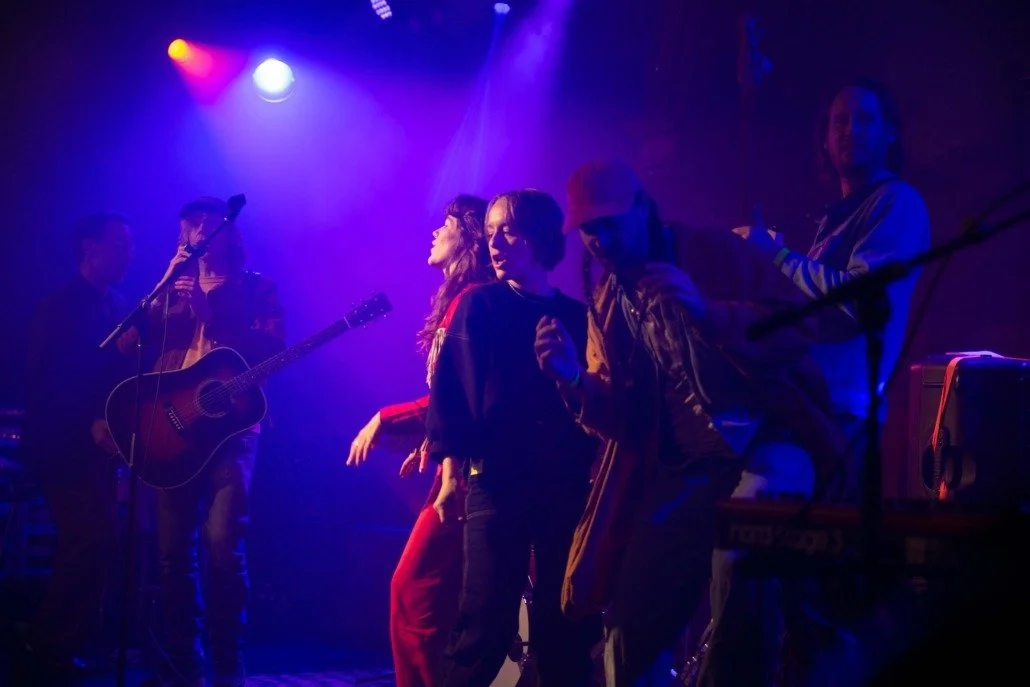 A live music performance with a band on stage under colorful lights, featuring a guitarist, singers, and possibly other musicians.