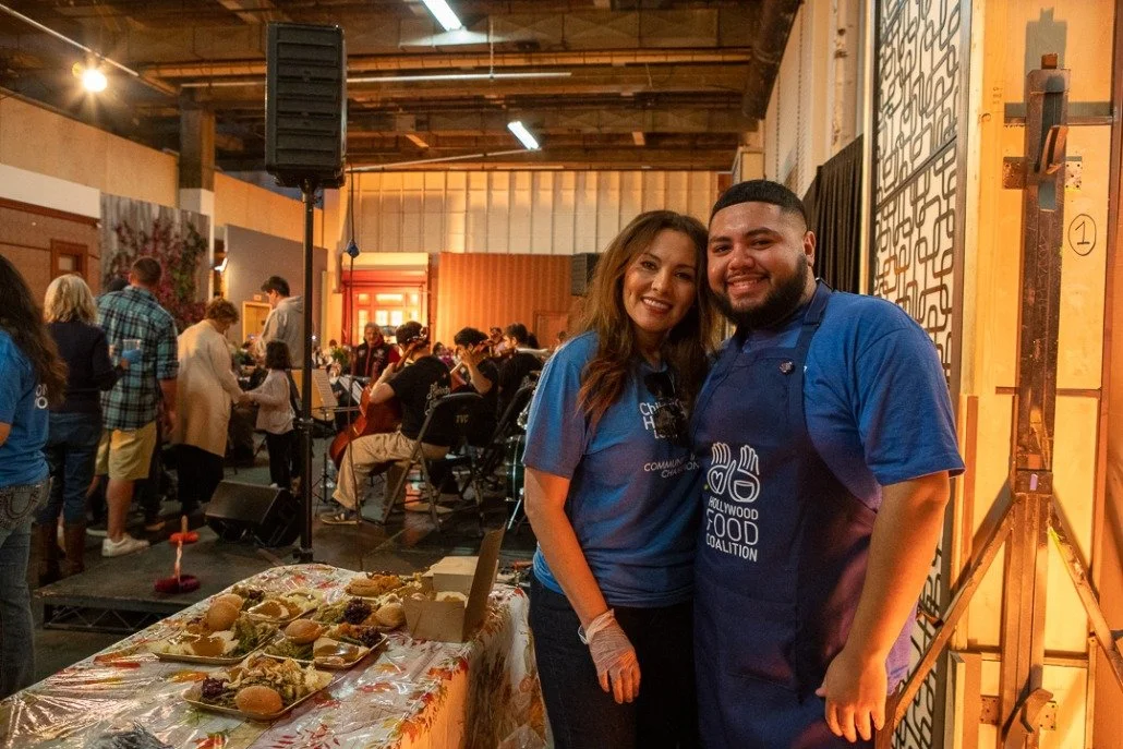 Two smiling people, a woman and a man, standing together at a food event, with a table of food in front of them and a crowd in the background.