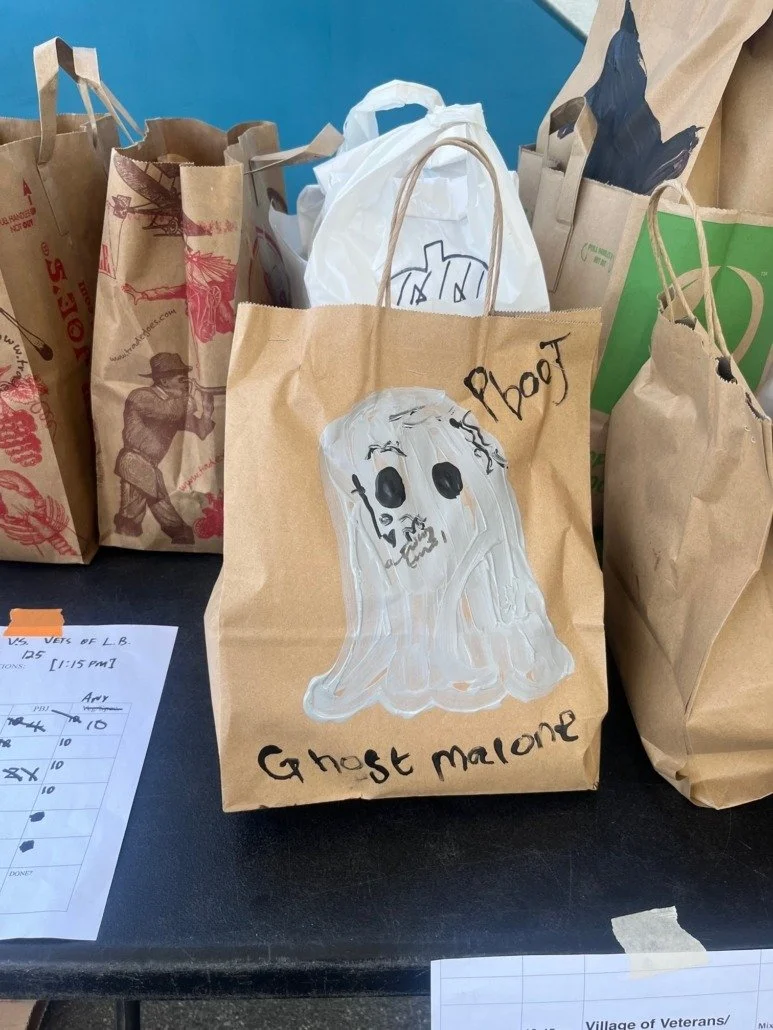Brown paper bag decorated with a ghost drawing, large black eyes, and the words 'Ghost Malone' and 'Ghost' written on it. Other paper bags and papers are visible on a table.