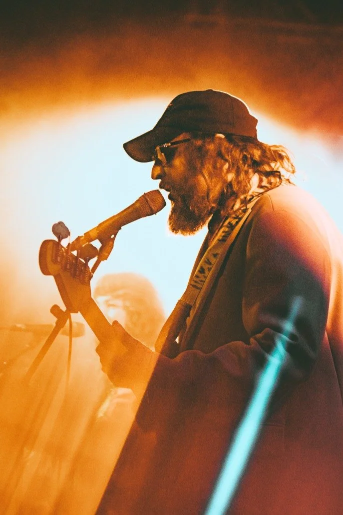 Musician singing into a microphone and playing an electric guitar on stage with warm lighting and a hazy background.