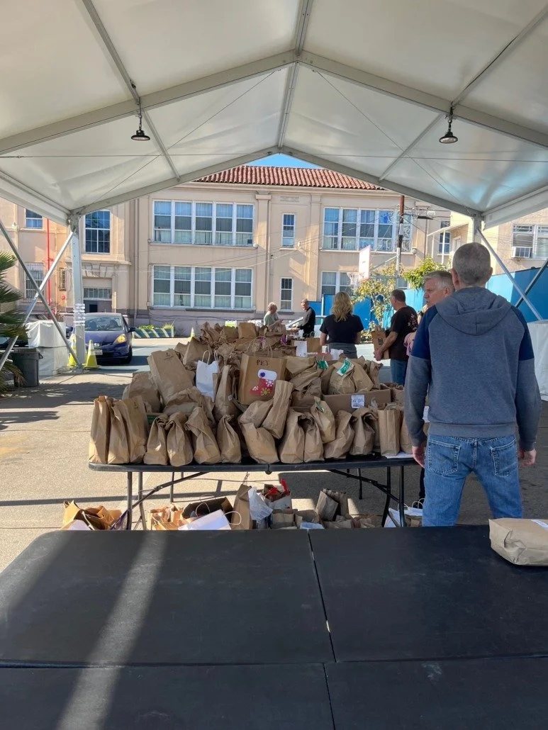 Table under a canopy with numerous paper gift bags, some with bows and tags, set up for an event.