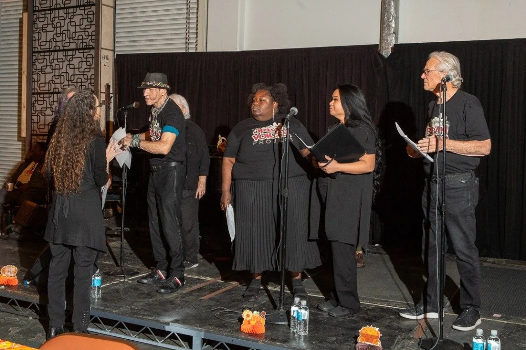 Six people standing on a stage with black curtains, participating in a discussion or presentation. One woman is speaking into a microphone, while others hold papers or folders. There are water bottles and small pumpkin decorations on the stage.