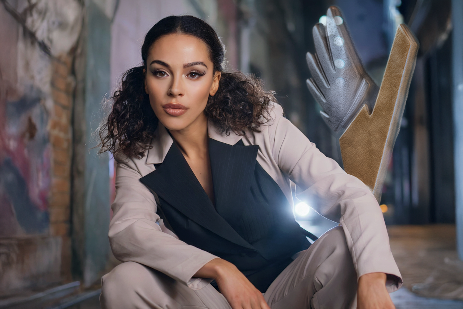 A woman with dark wavy hair and tan skin wearing a beige blazer and black shirt, sitting on the floor with her legs crossed, in an industrial-style space with exposed brick and wall art, and a large claw hand prop with metallic fingers next to her.