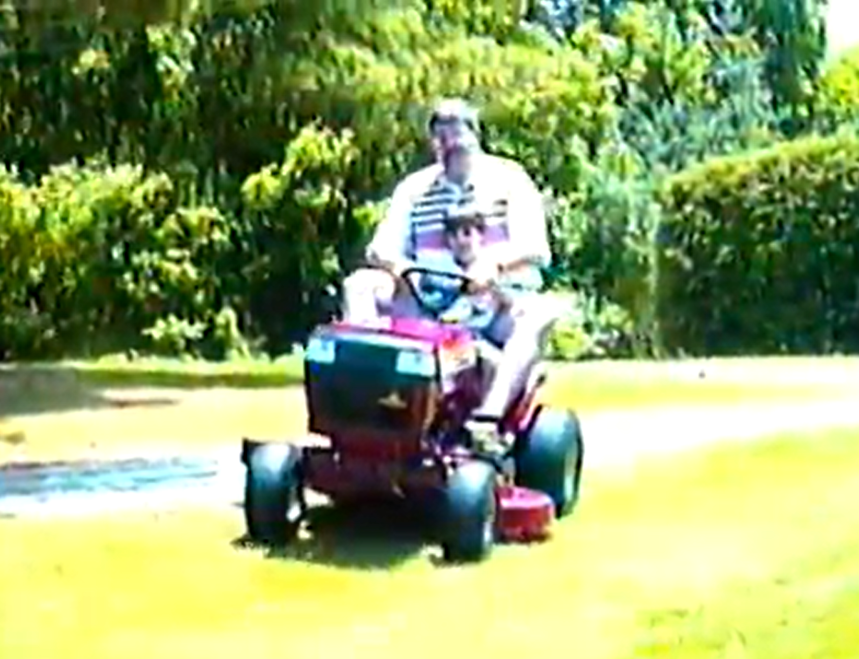 A man riding a red riding mower on a grassy lawn, with dense green bushes in the background.
