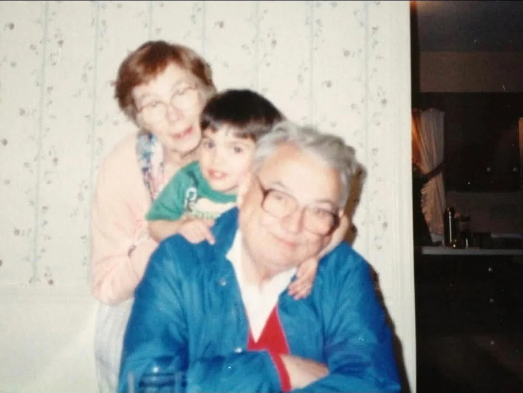 A family of three posing together indoors, including an elderly man with glasses, a woman with short red hair, and a young boy with dark hair. They are smiling and leaning on each other.
