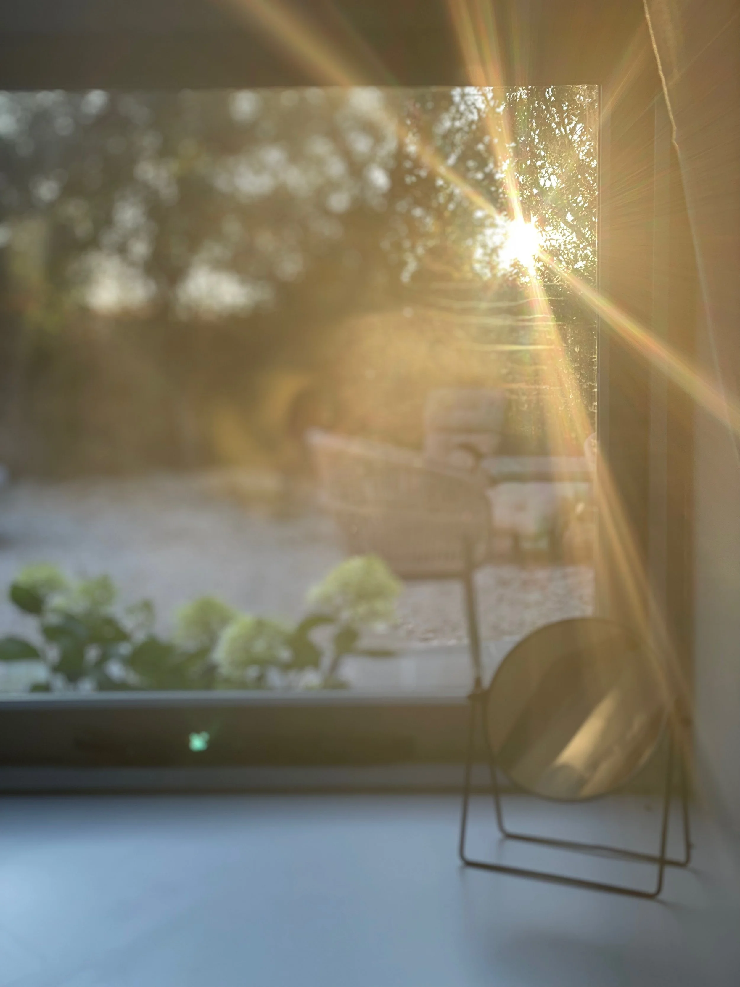 Sunlight shining through a window onto a windowsill with a small plant, a round mirror, and some outdoor chairs visible outside.