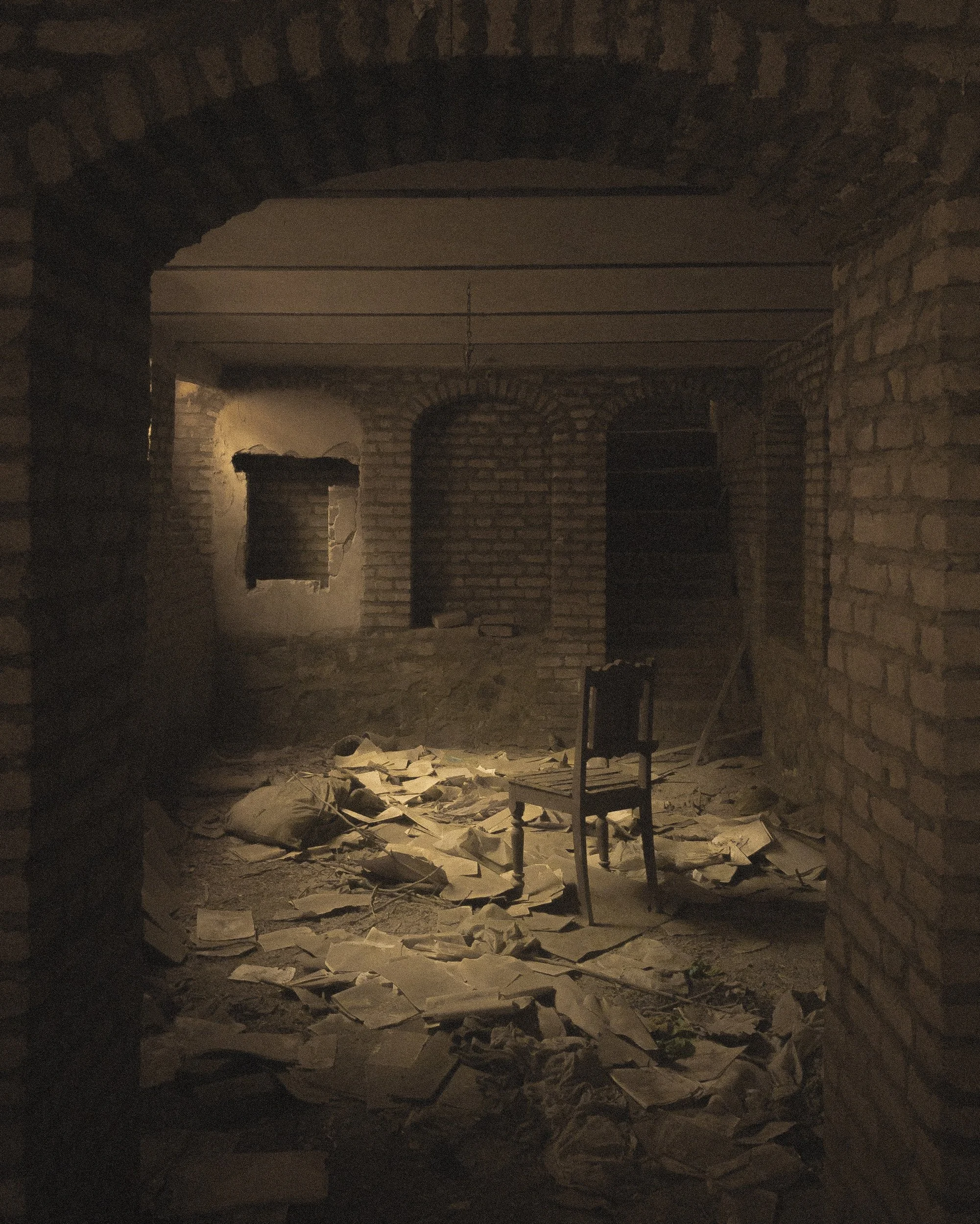 An abandoned, dimly lit basement or cellar with exposed brick walls, scattered papers and debris on the floor, an empty wooden chair, and a small window with a broken frame.