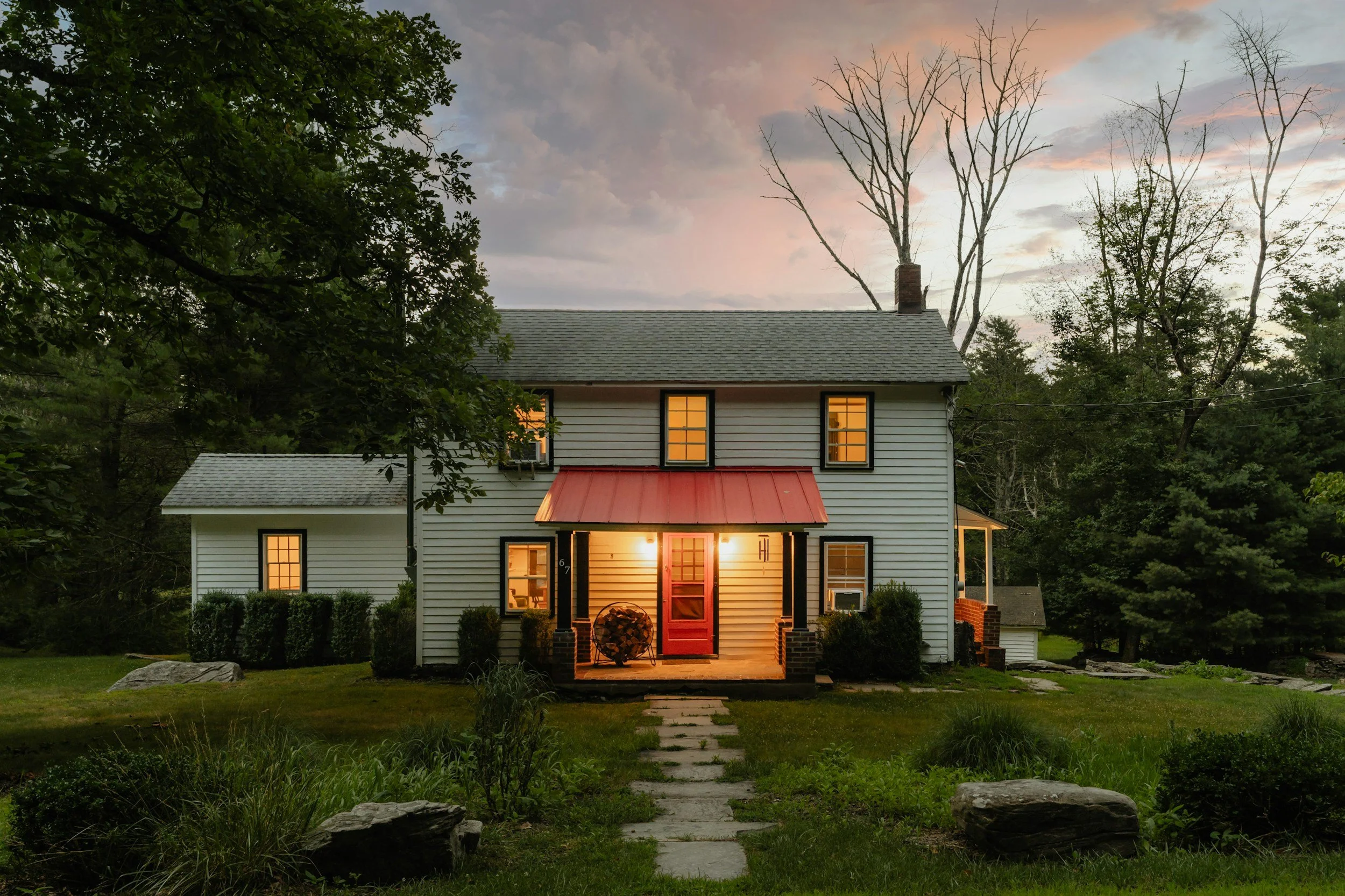 A two-story white house with black window frames and a red front door with lights on inside, illuminated at sunset, surrounded by trees and a grassy yard with stone pathway.