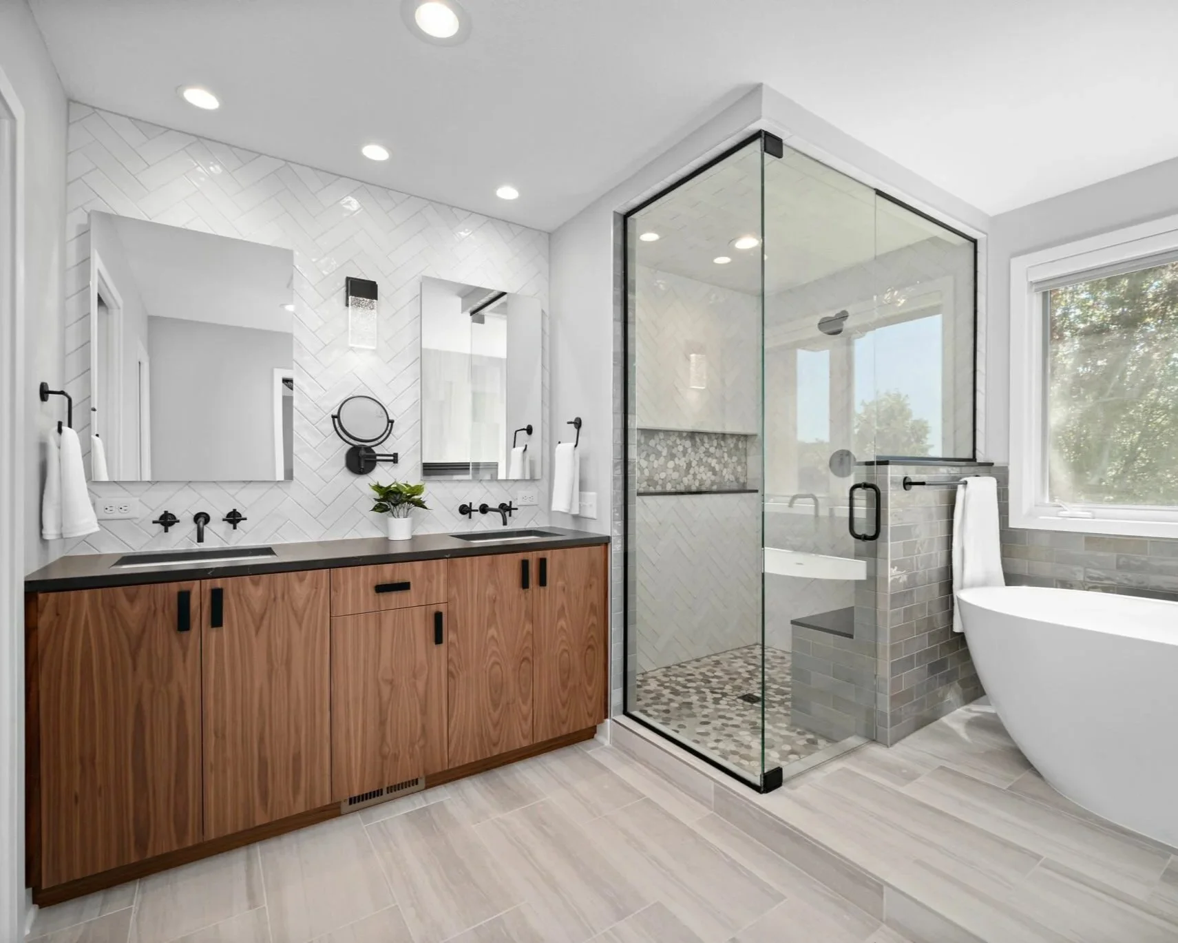 A modern bathroom with a wooden vanity, two sinks, and black fixtures. There are two mirrors, a potted plant, and a wall-mounted magnifying mirror. A glass-enclosed shower with patterned tile flooring and a bench, along with a freestanding bathtub beside a window, complete the space.