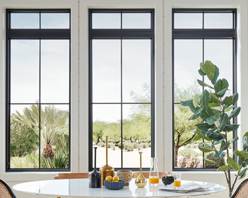 Interior dining area with three large windows showing a desert landscape with trees and palms outside; a white table with decorative vases, candles, and a glass of orange juice, with a tall potted plant nearby.