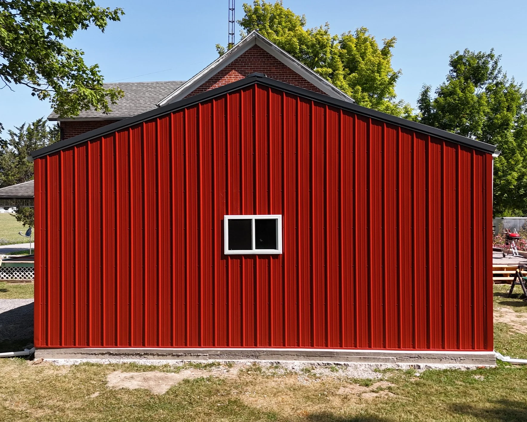 Red metal building with a small window, situated on a grassy area with trees and a house in the background.