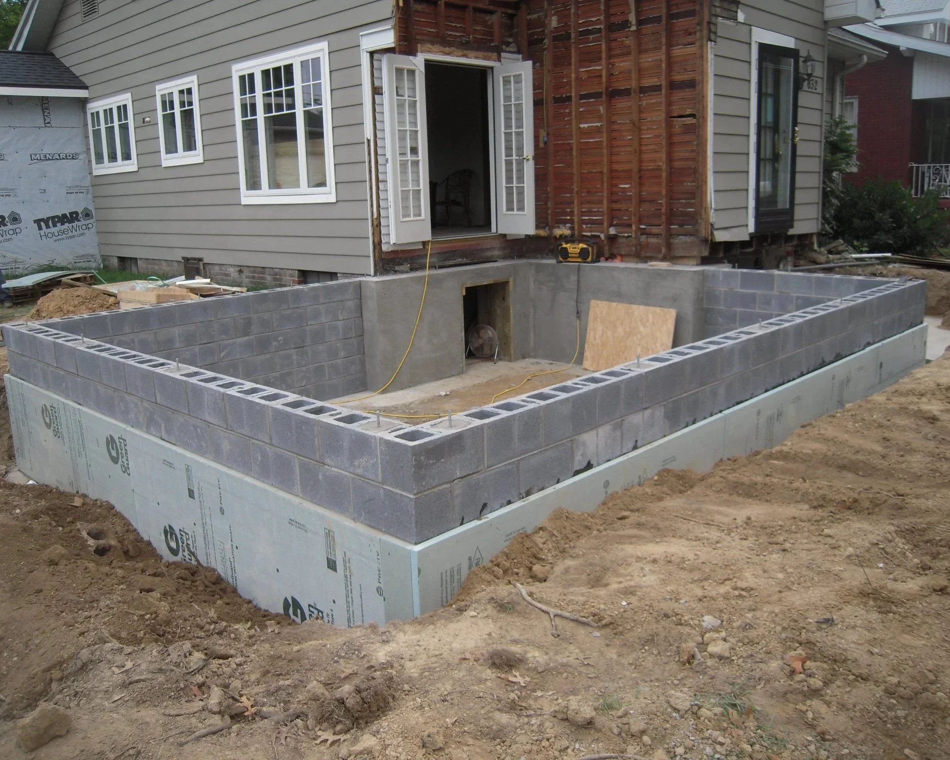 Construction site of a backyard pool with concrete block walls built in a rectangular shape next to a house under renovation, with exposed wooden framing and an open door.