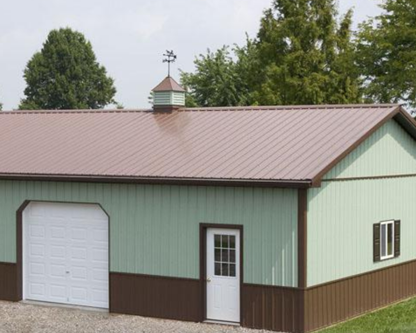 A light green barn with a brown lower section and a brown metal roof, featuring a white garage door, a small white door, and a window, surrounded by trees.