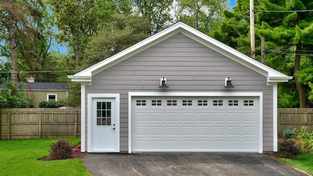 Gray garage with white door and side door, surrounded by green lawn and trees, with a wooden fence in the background.
