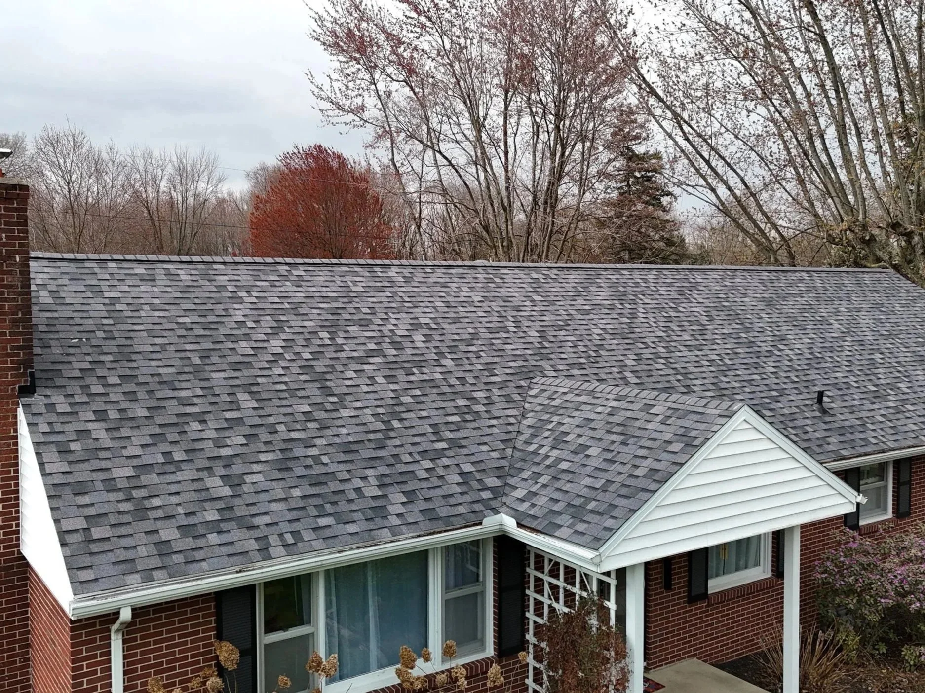 A residential house with a gray shingle roof, brick walls, white porch and window trim, surrounded by trees with autumn foliage.