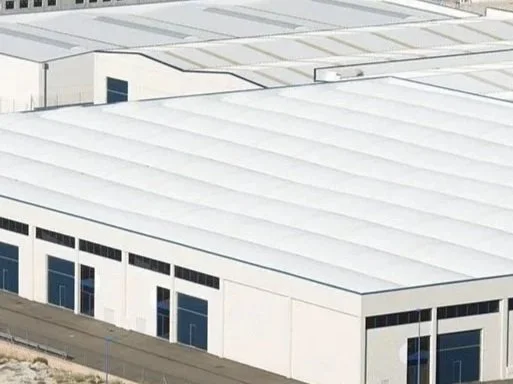 A large industrial warehouse with a white roof and white walls, featuring several blue doors.