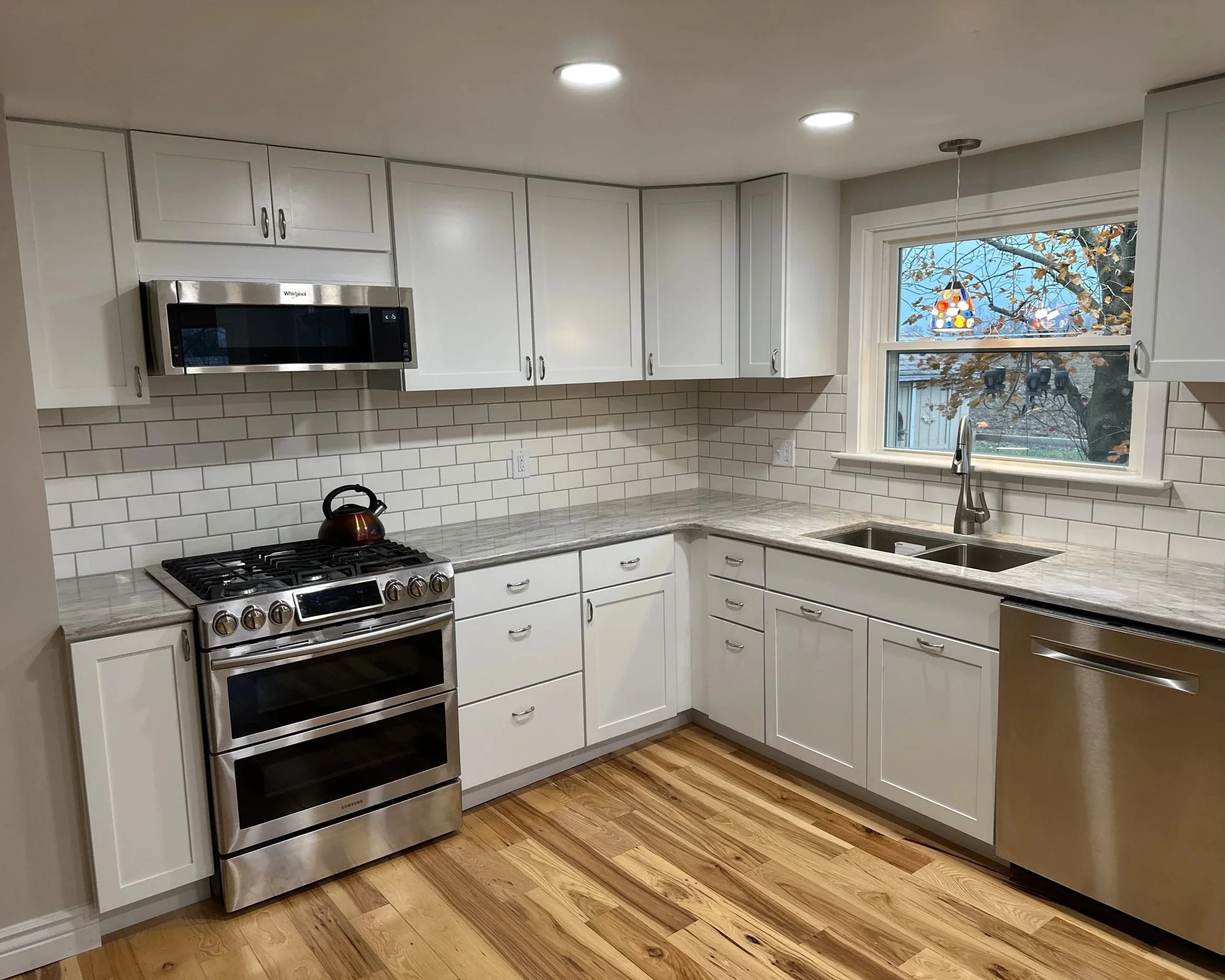 Modern kitchen with white cabinets, a stainless steel oven, microwave, and dishwasher, granite countertops, white subway tile backsplash, wood flooring, and a window with a view of a tree with autumn leaves.