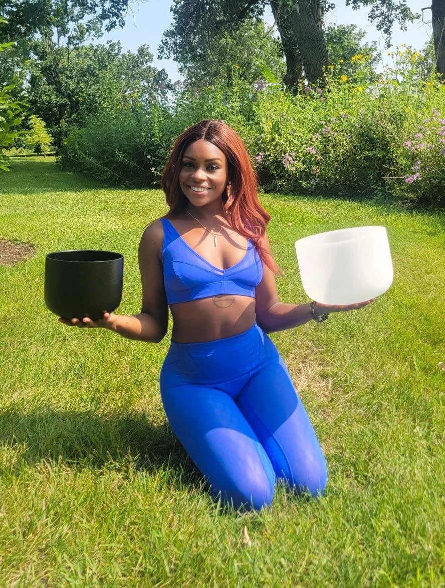 A woman with red hair kneeling on grass outdoors, holding a black singing bowl in her right hand and a white singing bowl in her left hand, smiling at the camera, surrounded by greenery and trees.