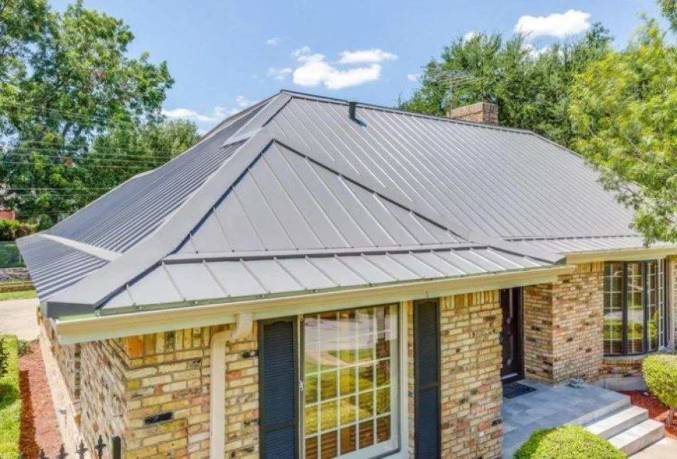 A house with a gray metal roof, brick exterior, and a small front porch with steps, surrounded by greenery and trees under a blue sky.