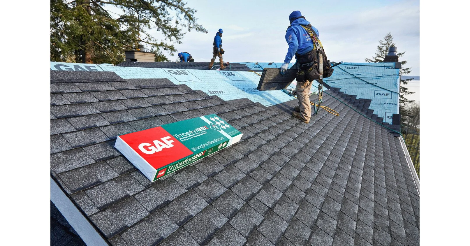 Roofers installing asphalt shingles on a house roof, with a GAF Timberline UHD shingle box in the foreground and workers wearing blue uniforms and safety harnesses.