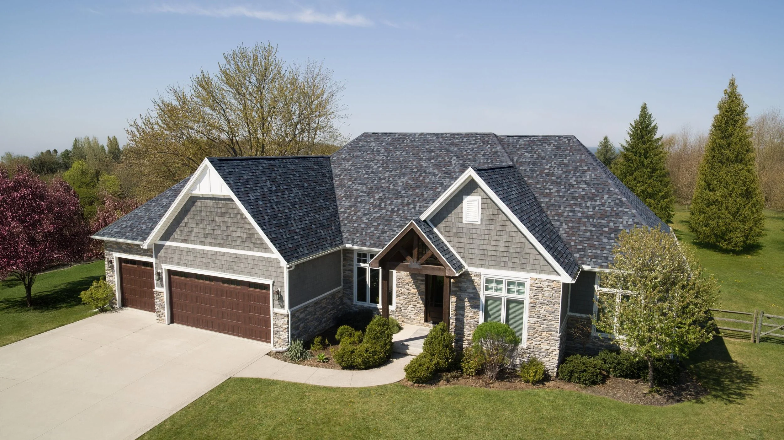 A modern house with a stone and wood exterior, a brown garage door, and a shingled roof, surrounded by trees and a green lawn.