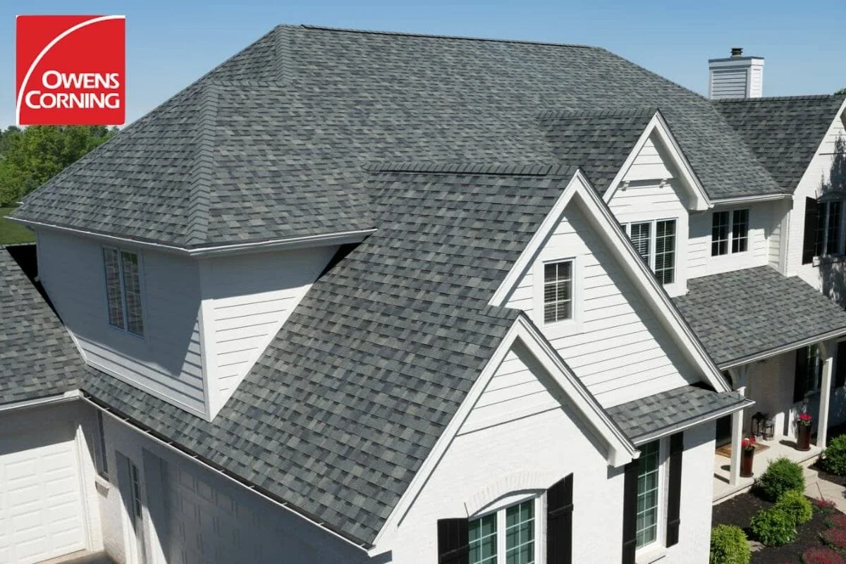 A large house with a gray asphalt shingle roof, white siding, and multiple gables. The front yard has bushes and potted plants.