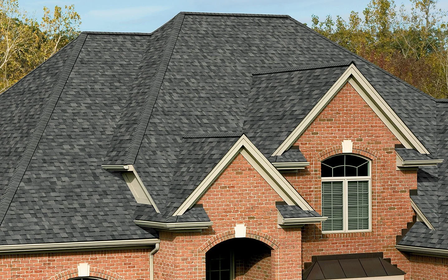 Close-up of a house roof with dark gray shingles, brick exterior walls, and a large arched window with shutters in the front.