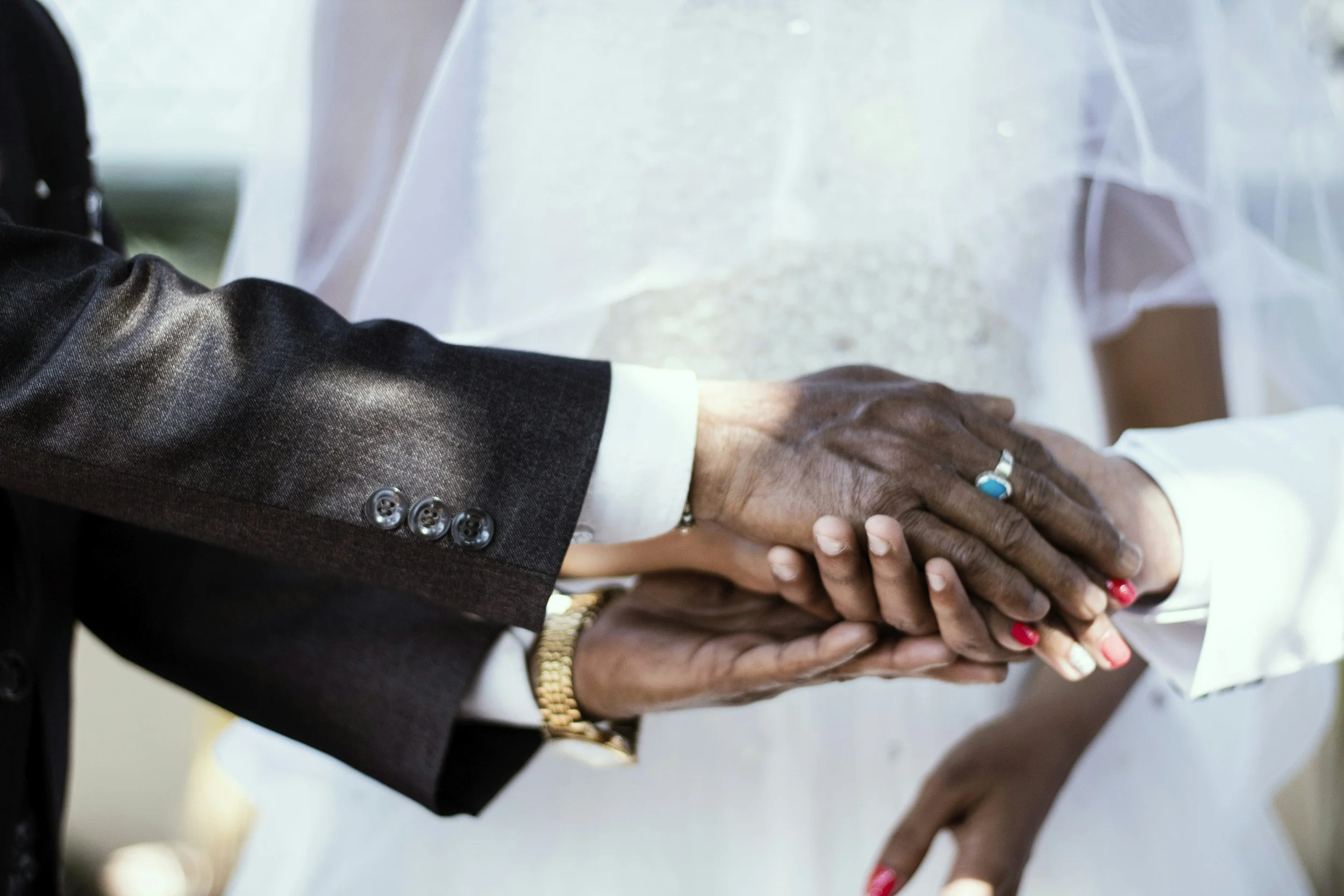 Close-up of two people's hands gently holding each other, one wearing a dark suit with buttons on the sleeve and a white shirt, and the other wearing multiple rings and a bracelet. The background appears to be a light-colored surface.