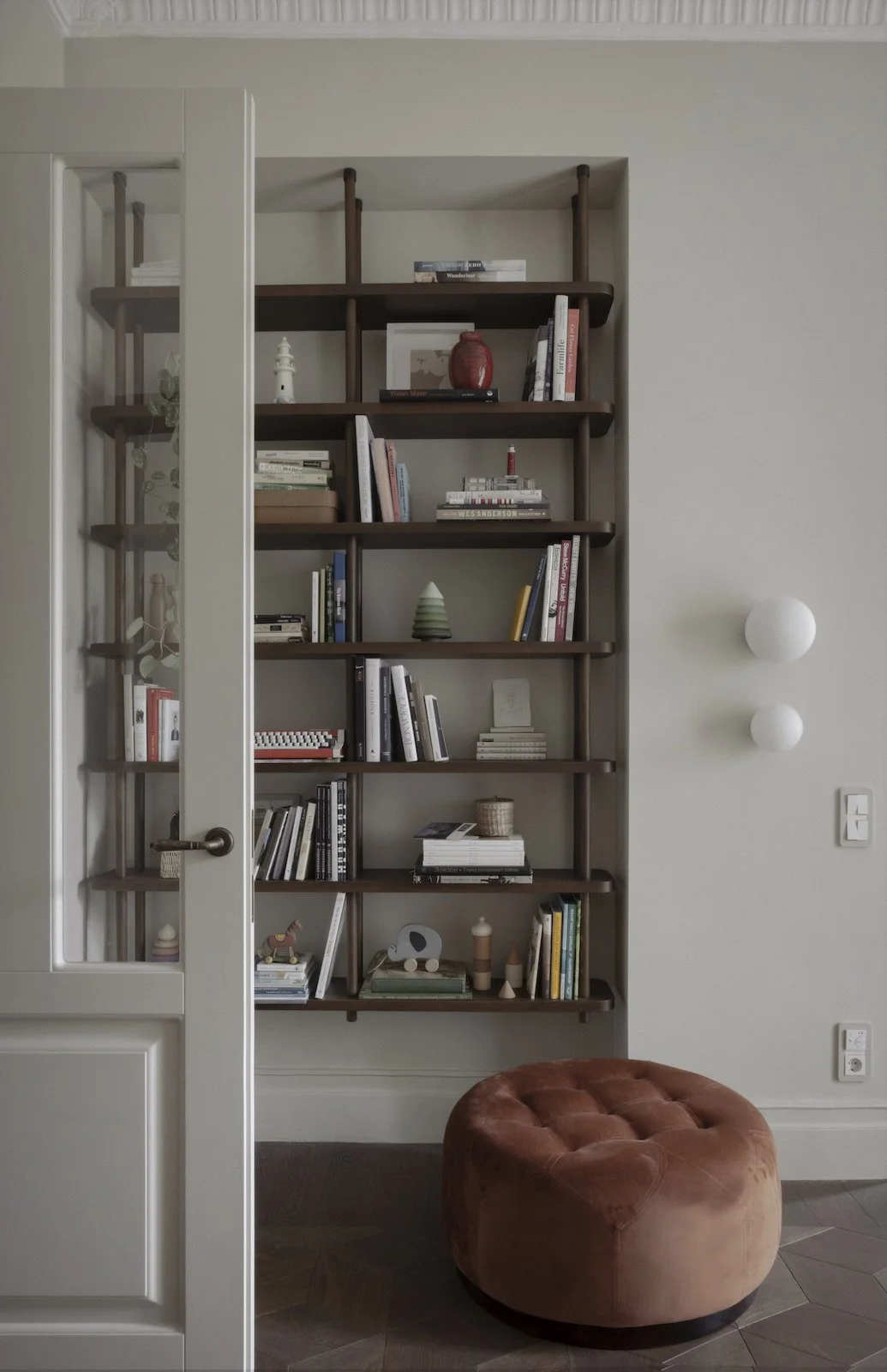 Living area of Goldfish Apartments by Jenny Ly Studio — built-in shelving, soft velvet pouf, and muted wall palette.
