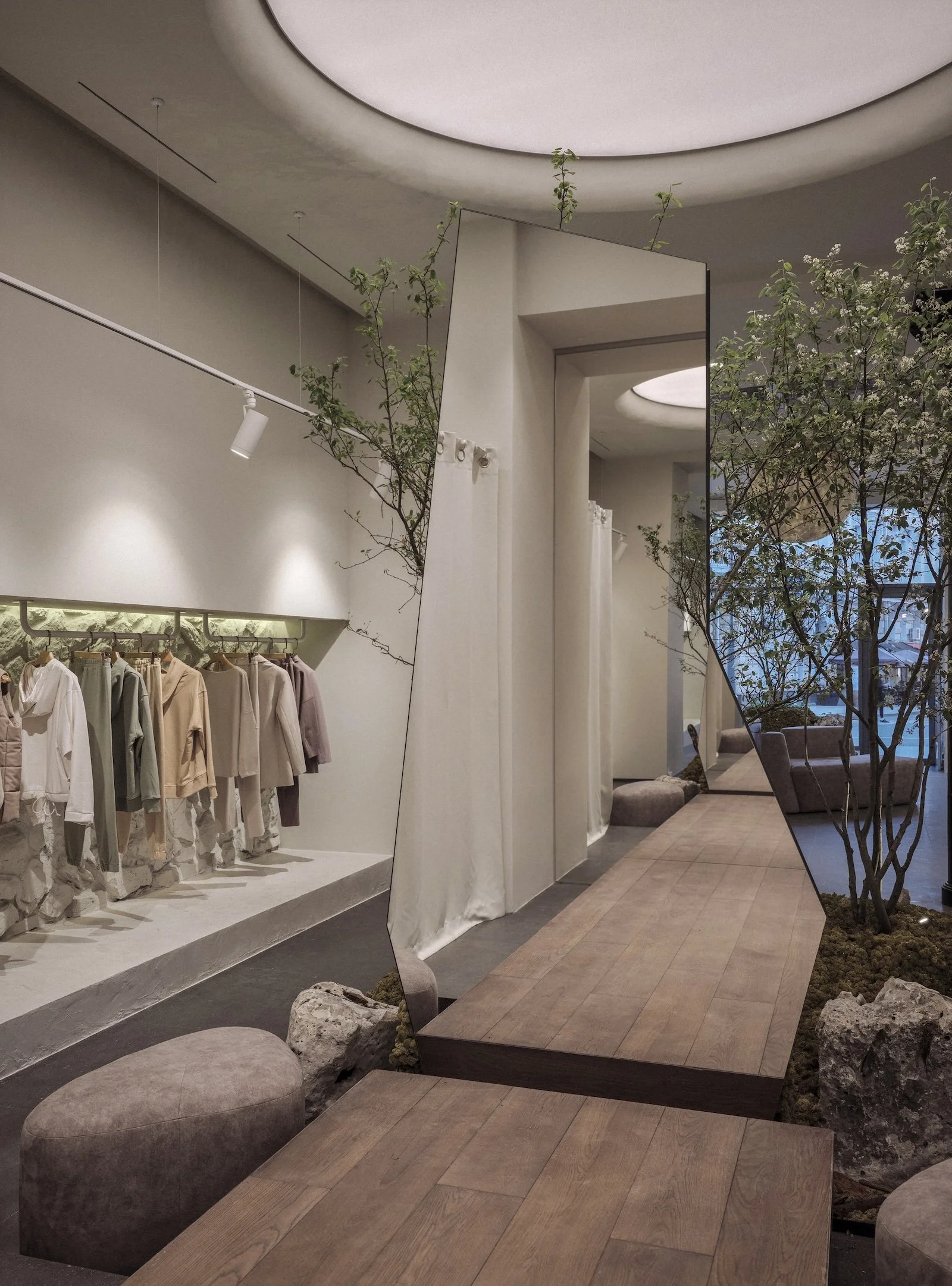 Fitting area in a clothing showroom with elevated podium, large mirror, soft ceiling light, and a tree visible behind the glass - interior by Jenny Ly Studio.