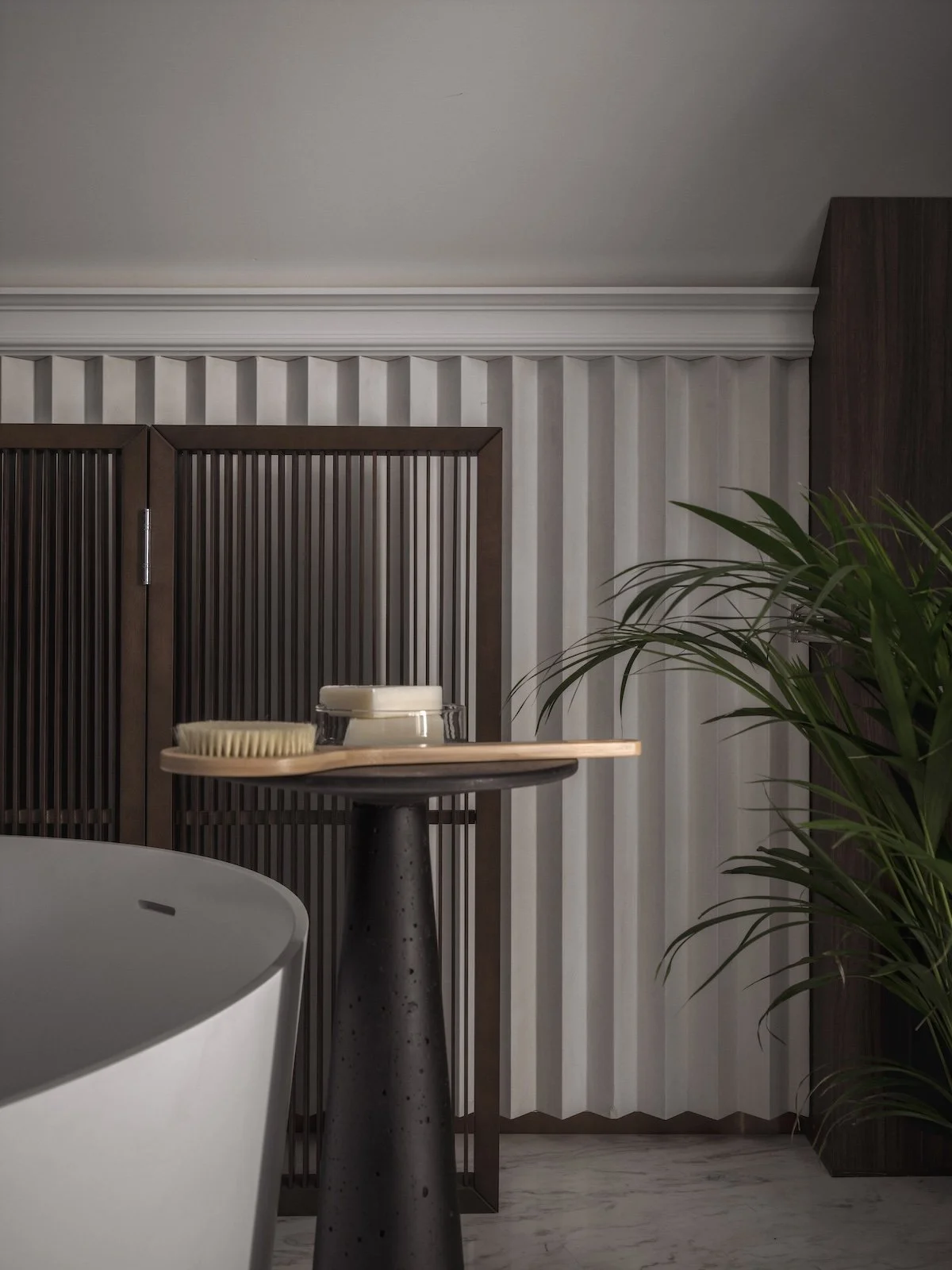Interior of a modern room with a side table holding a brush, glass container, and a container with a lid, a white bathtub in the foreground, a large green plant to the right, and wooden decorative panels on the wall.
