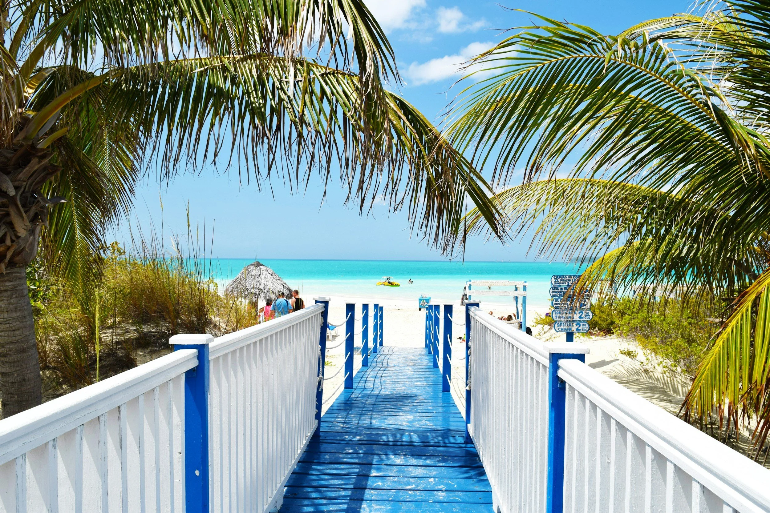 Beach entrance with white and blue railing leading to sandy shore with turquoise water, palm trees, and a thatched umbrella.