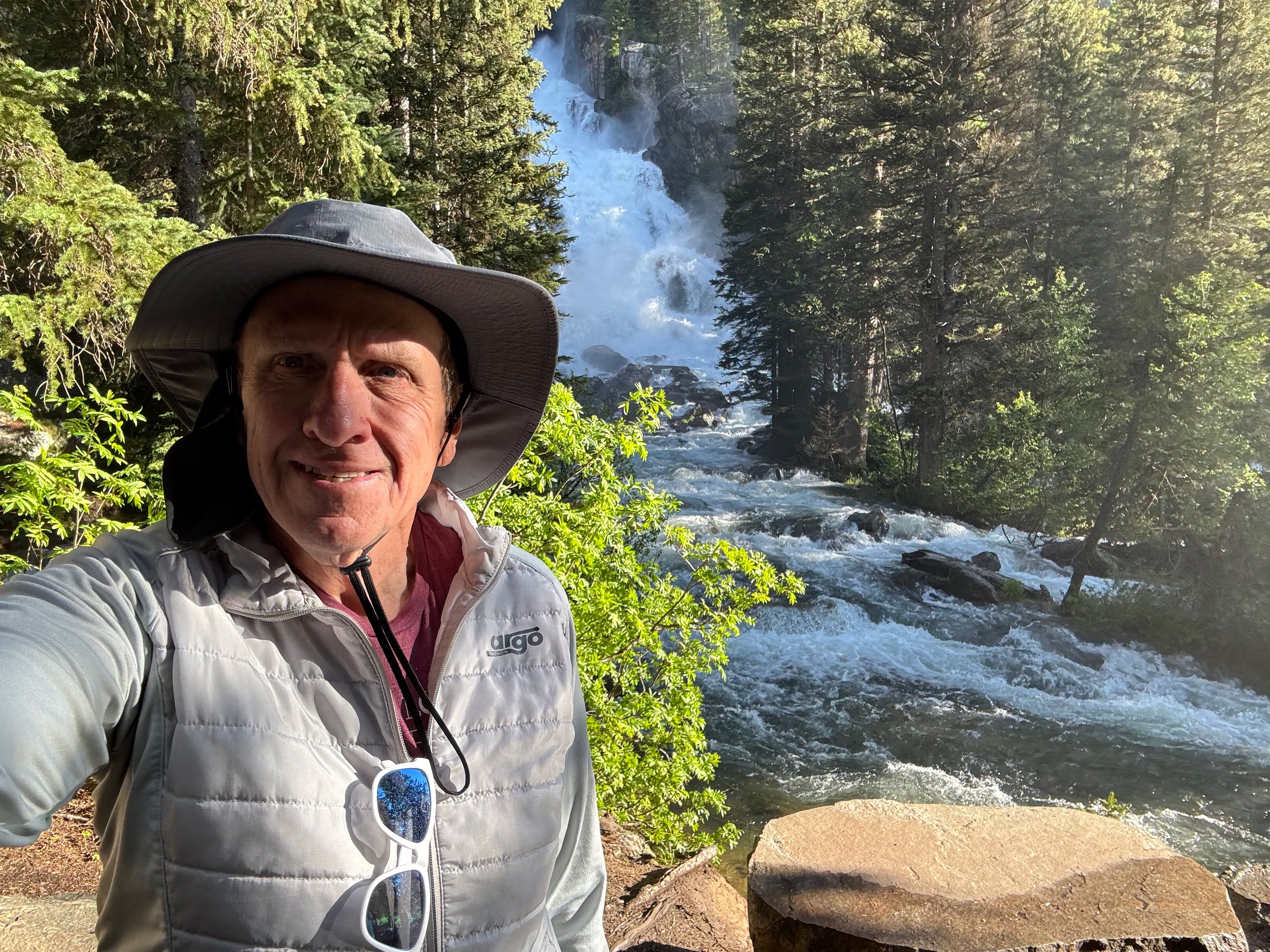 A picture of Eric hiking in front of a waterfall. The trees are lush and bright green!