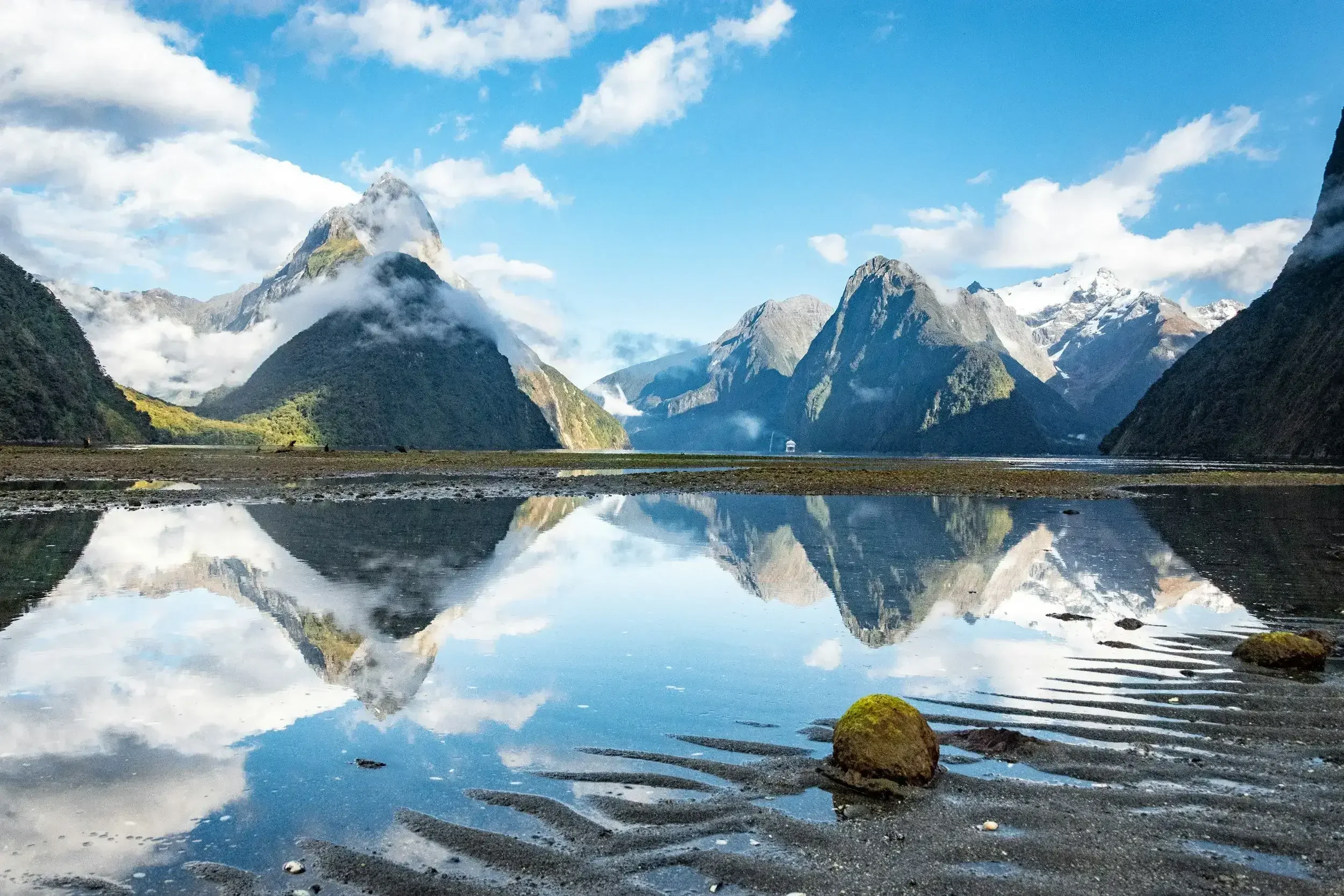 Snow-capped mountains and lush green hills reflected in a calm body of water along a rocky shoreline under a partly cloudy sky.