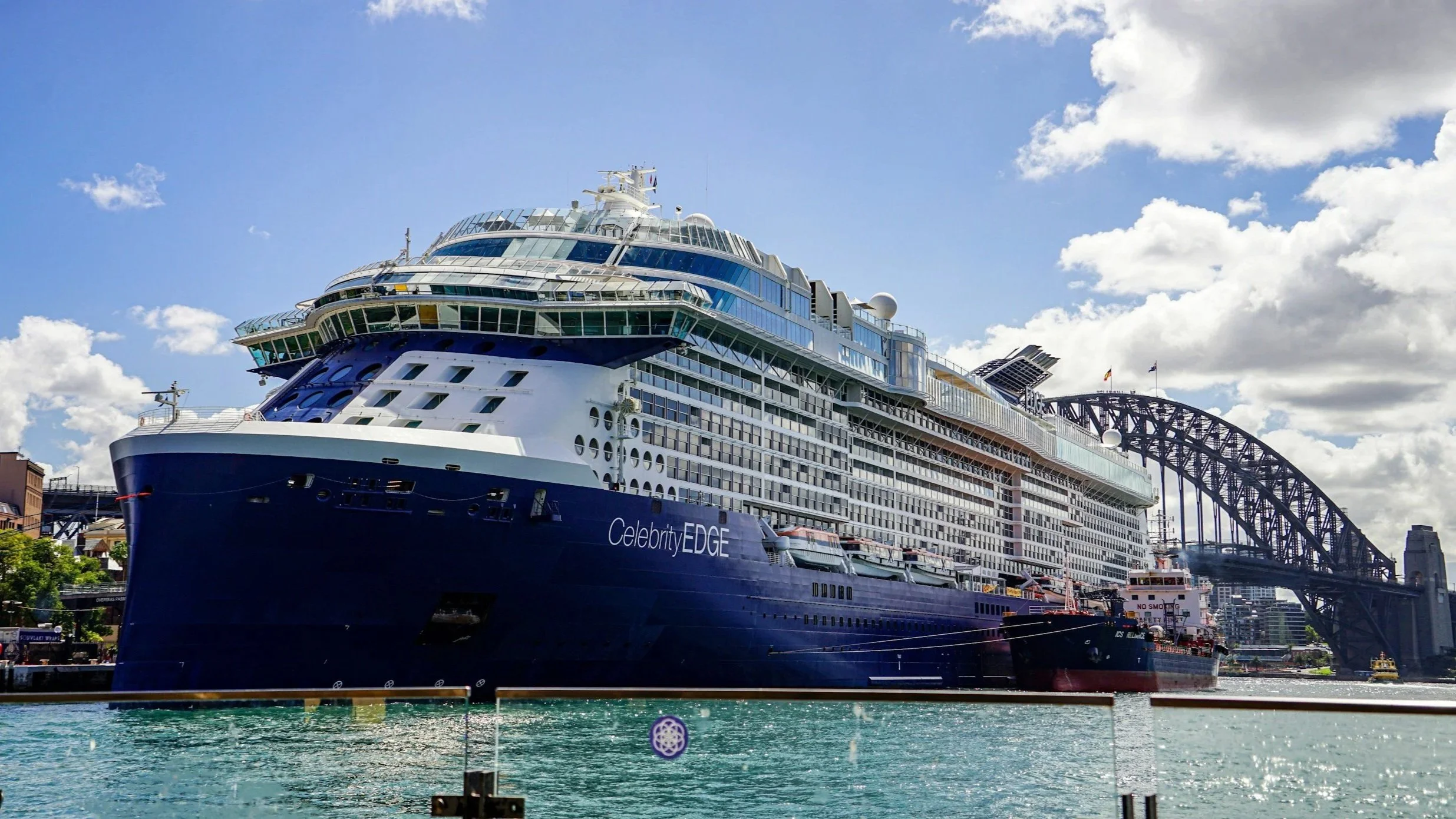 A large cruise ship named Celebrity Edge docked at a harbor with the Sydney Harbour Bridge in the background on a sunny day.