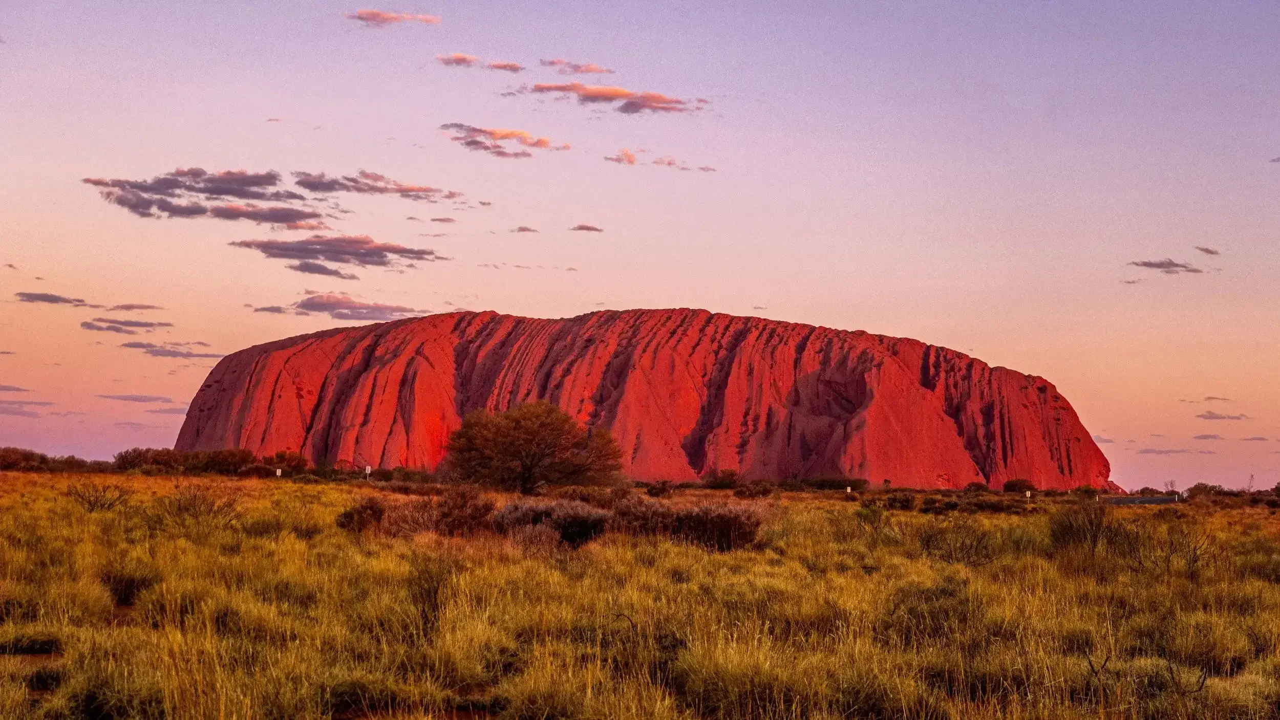 A large, reddish-brown rock formation, Uluru, is seen in a desert landscape during sunset, with a sky filled with a few scattered clouds.