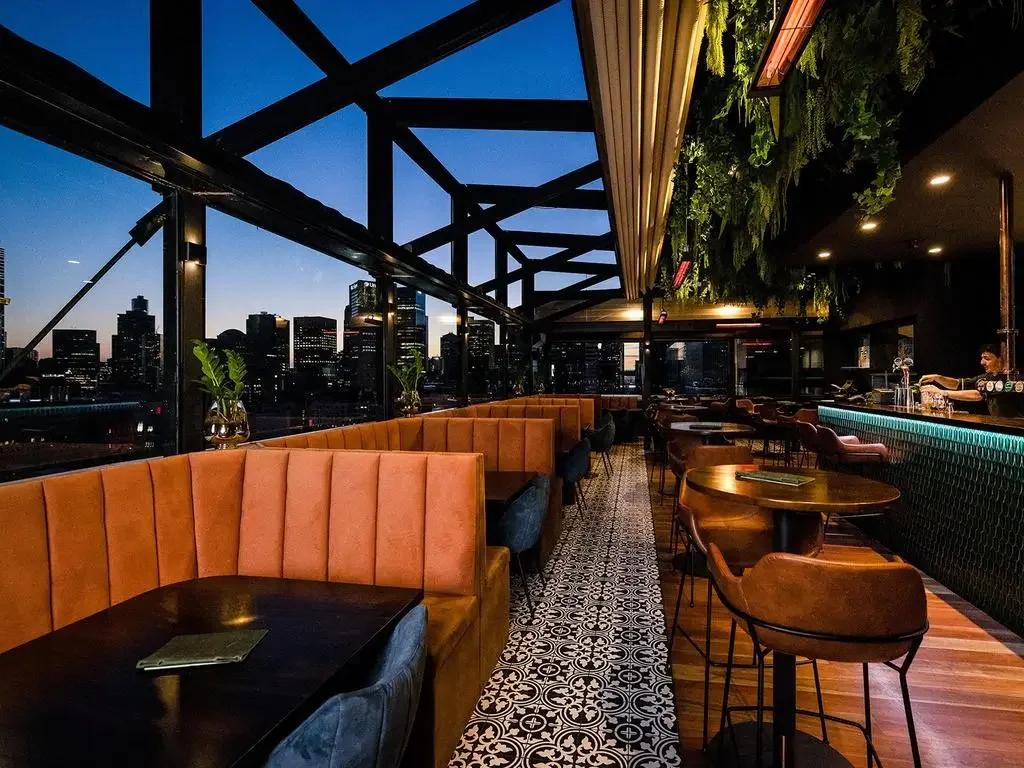 Interior of a modern rooftop bar or restaurant during sunset, with city skyline view. Features orange and dark blue upholstered seating, round wooden tables, decorative ceiling with greenery, patterned floor tiles, and a bar area with a bartender.