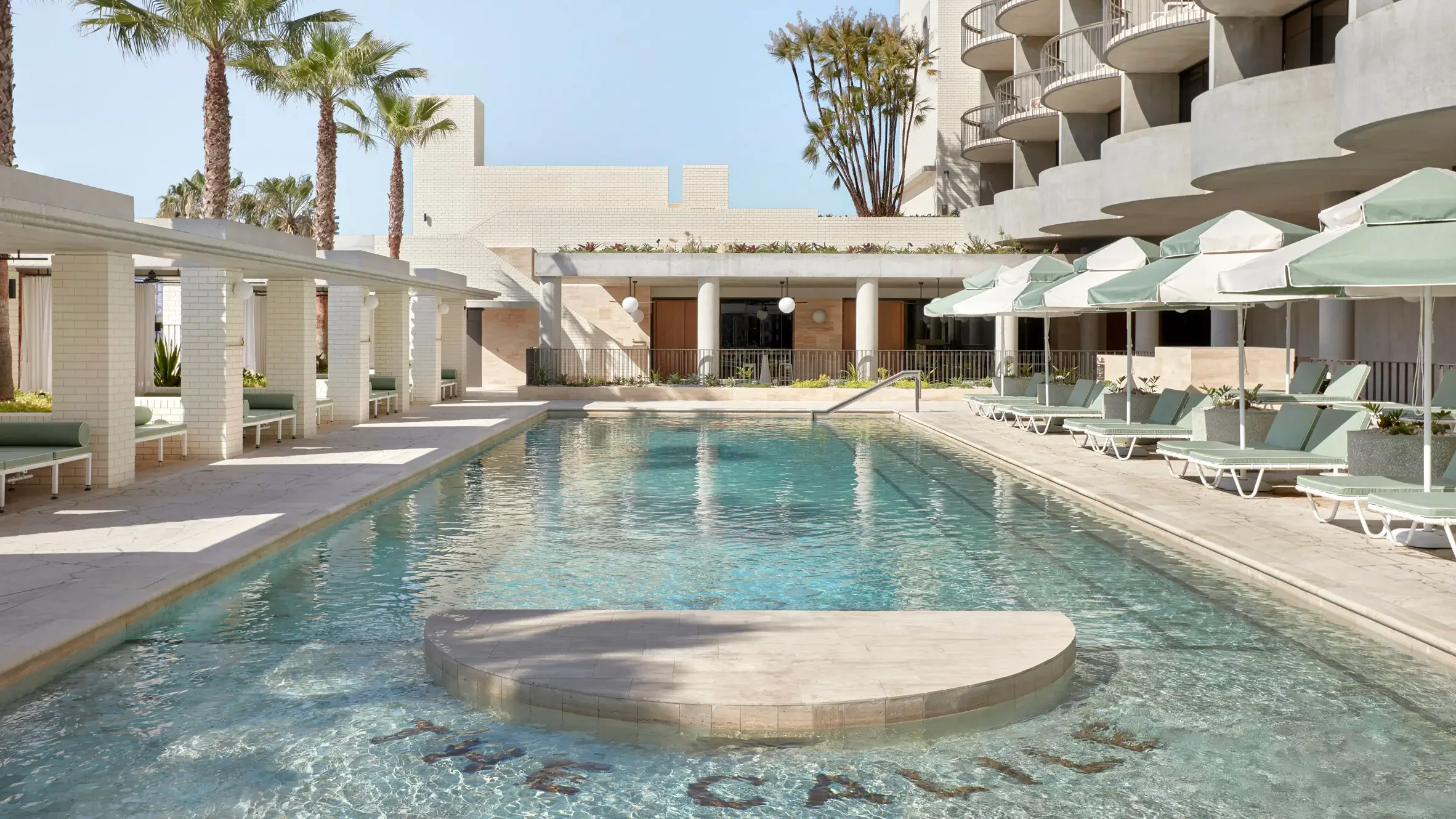 Empty outdoor swimming pool surrounded by white lounge chairs and umbrellas at a hotel, with palm trees and modern building architecture in the background.