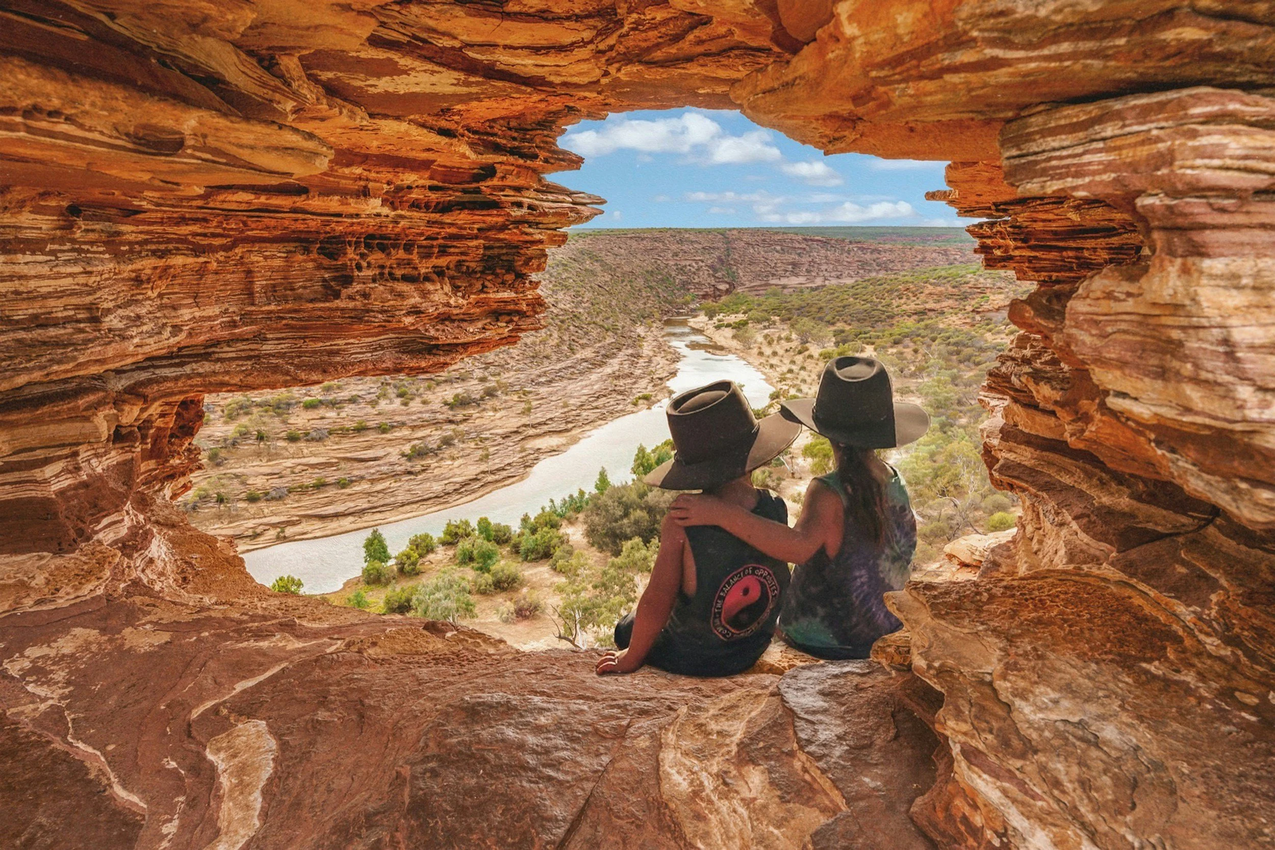 Two people wearing hats sitting inside a rock formation, overlooking a river and a vast desert landscape with sparse vegetation and a few trees, under a partly cloudy sky.