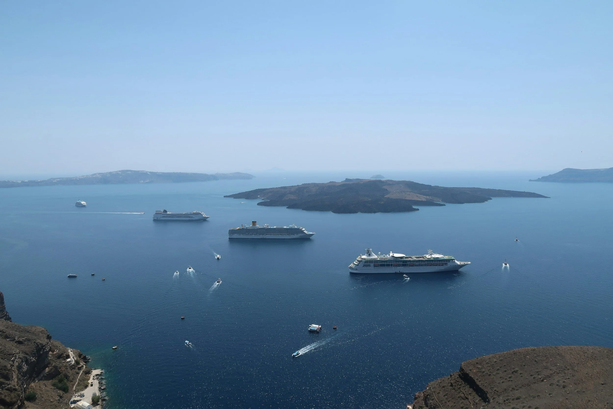 A photo of a body of water with multiple cruise ships and smaller boats, and landmasses in the distance, taken from a high vantage point.
