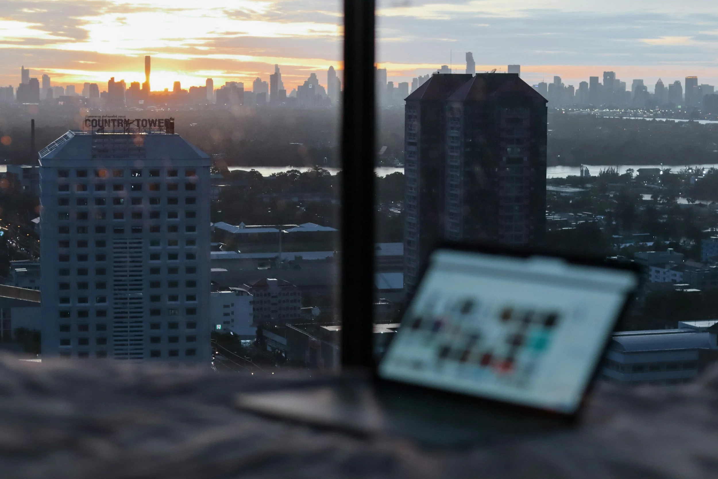 City skyline at sunset seen through a window with buildings, including a tall skyscraper, and a river in the background.