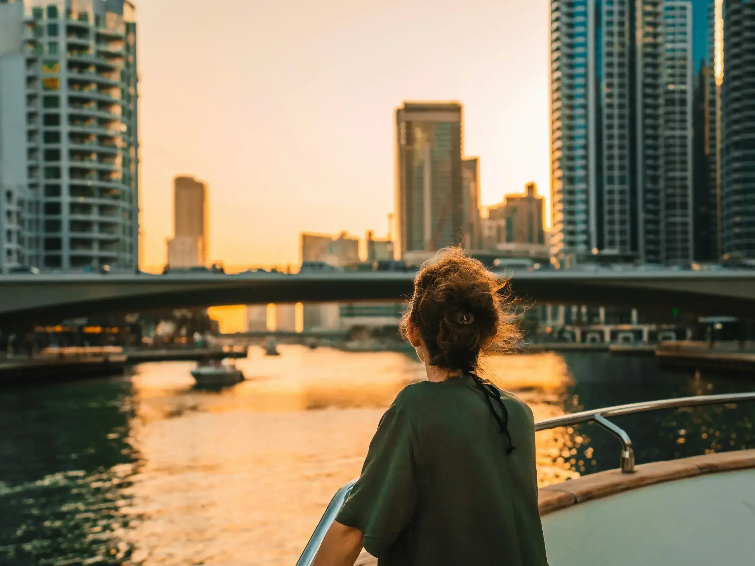 A person with curly hair tied back, wearing a dark green shirt, stands on a boat looking toward a cityscape with high-rise buildings along a river at sunset.