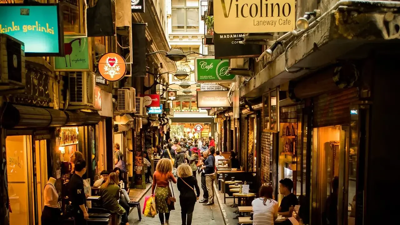 A busy alley with cafes and shops, many people sitting and walking, illuminated by warm lighting.
